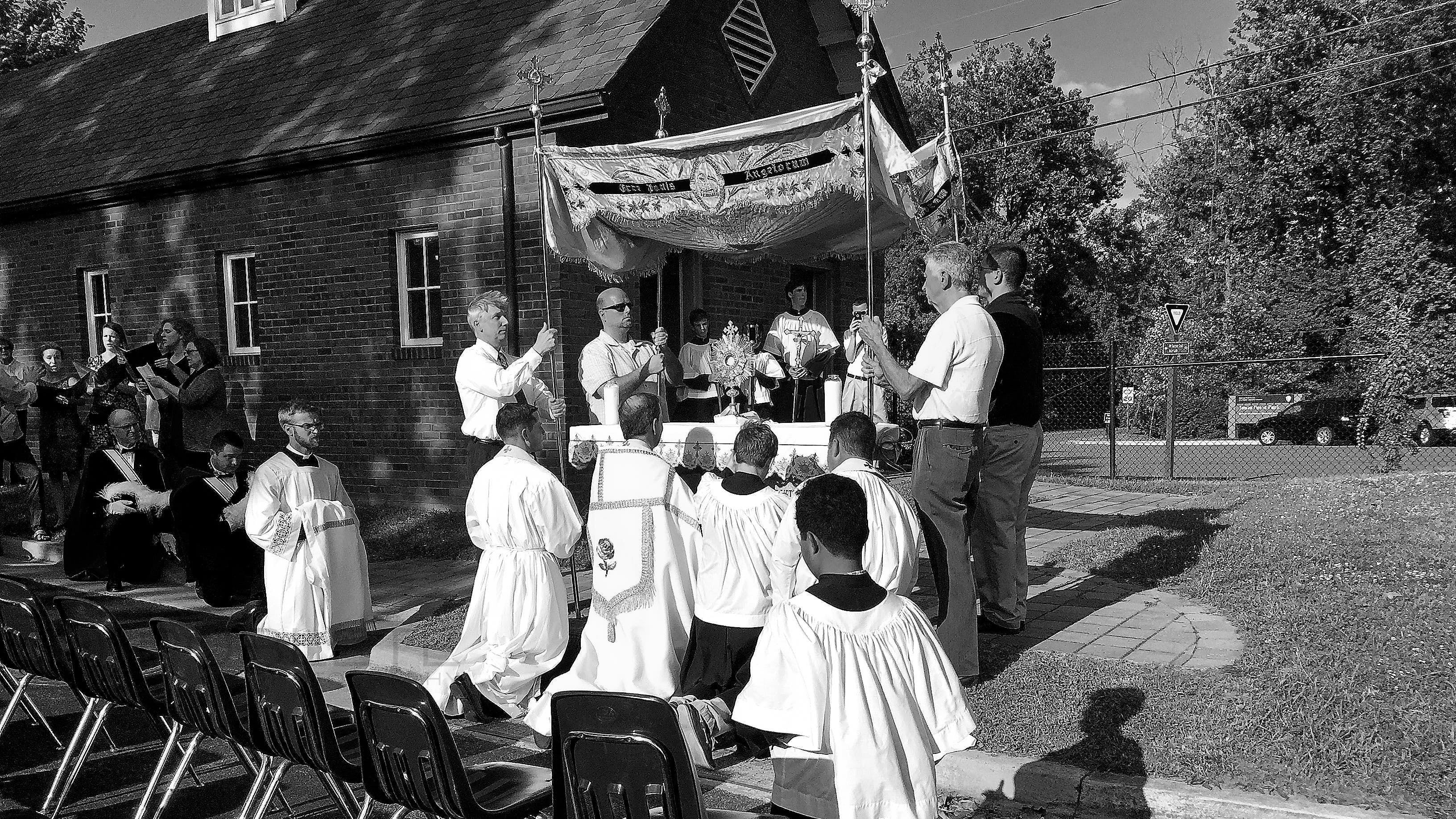 Clergy and altar servers gather outdoors for a ceremony beside a brick church.