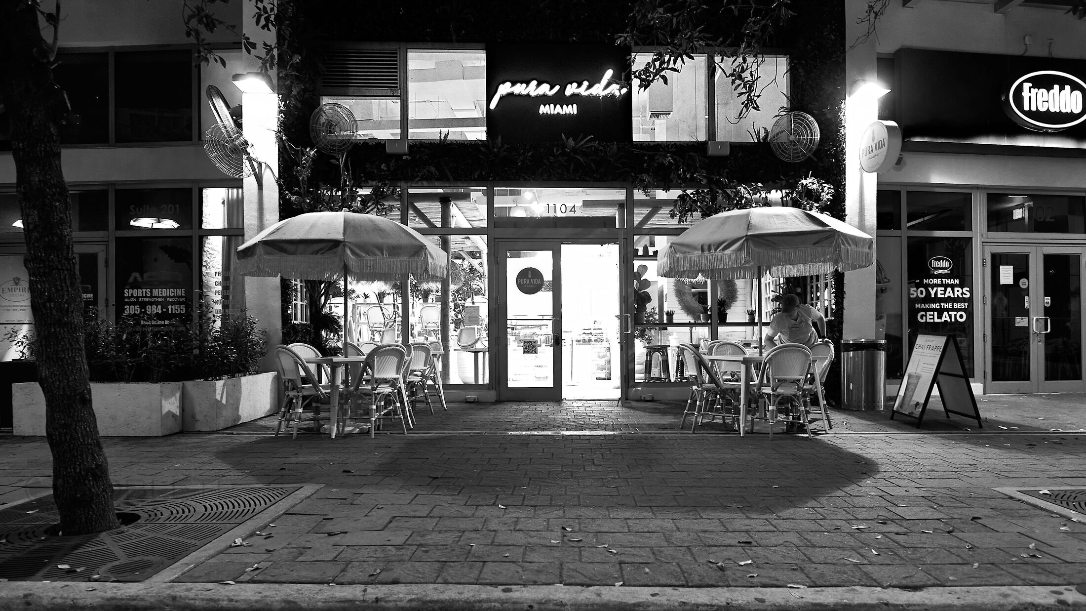 An illuminated café storefront and empty tables face a quiet street at dawn.