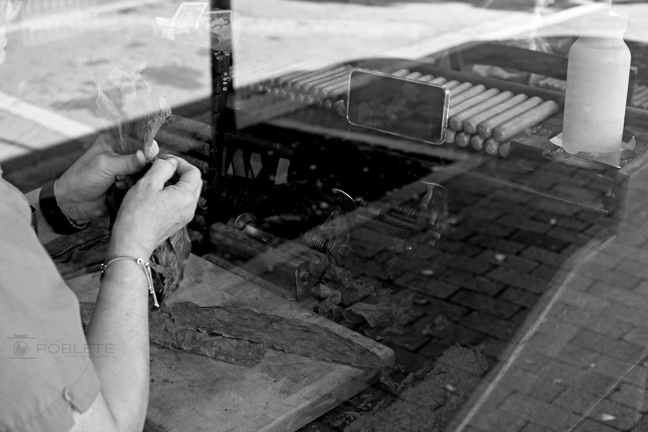 A cigar maker works with tobacco leaves behind a reflective shop window.