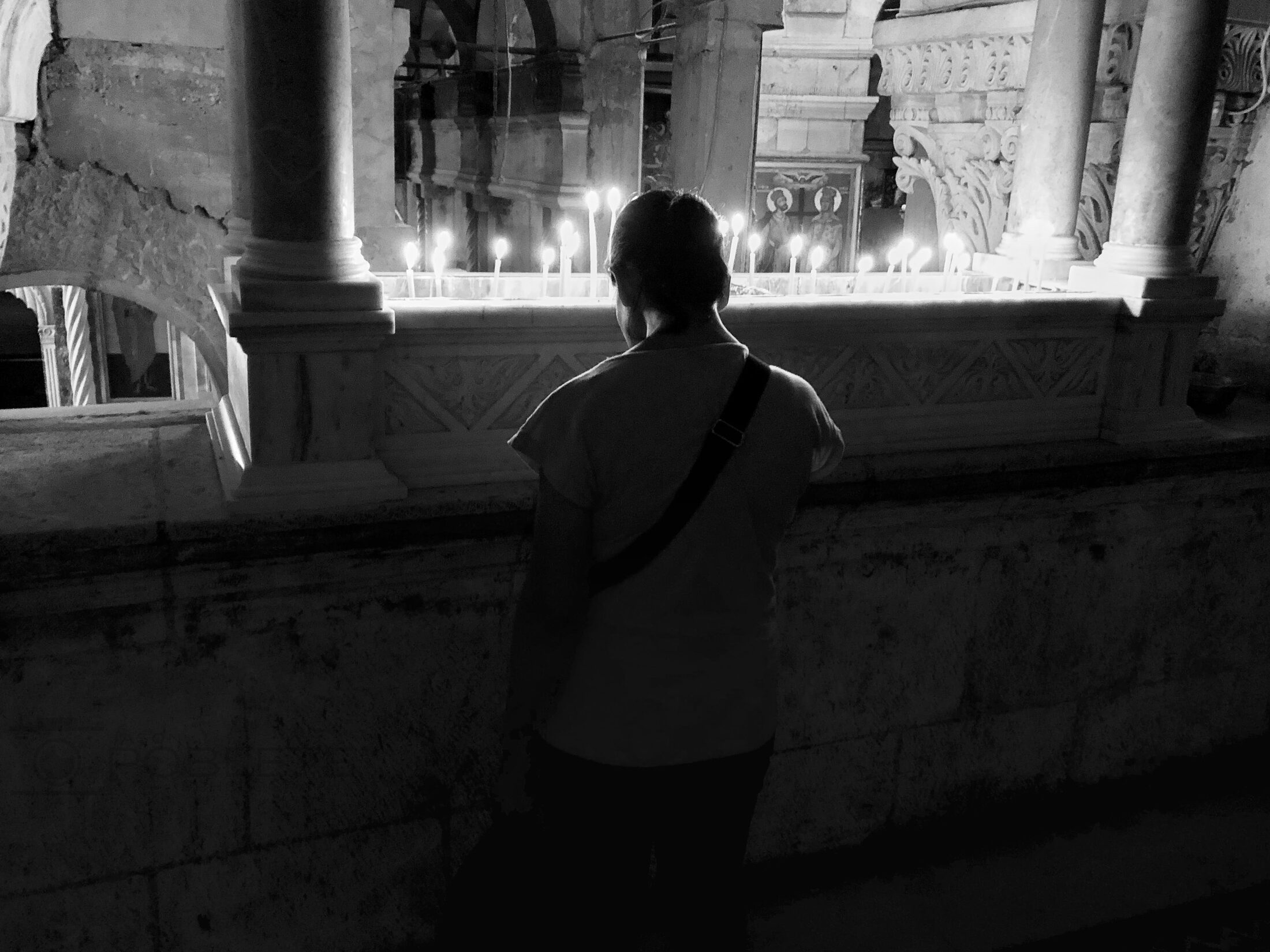 A woman stands in prayer before a row of candles in a stone chapel.
