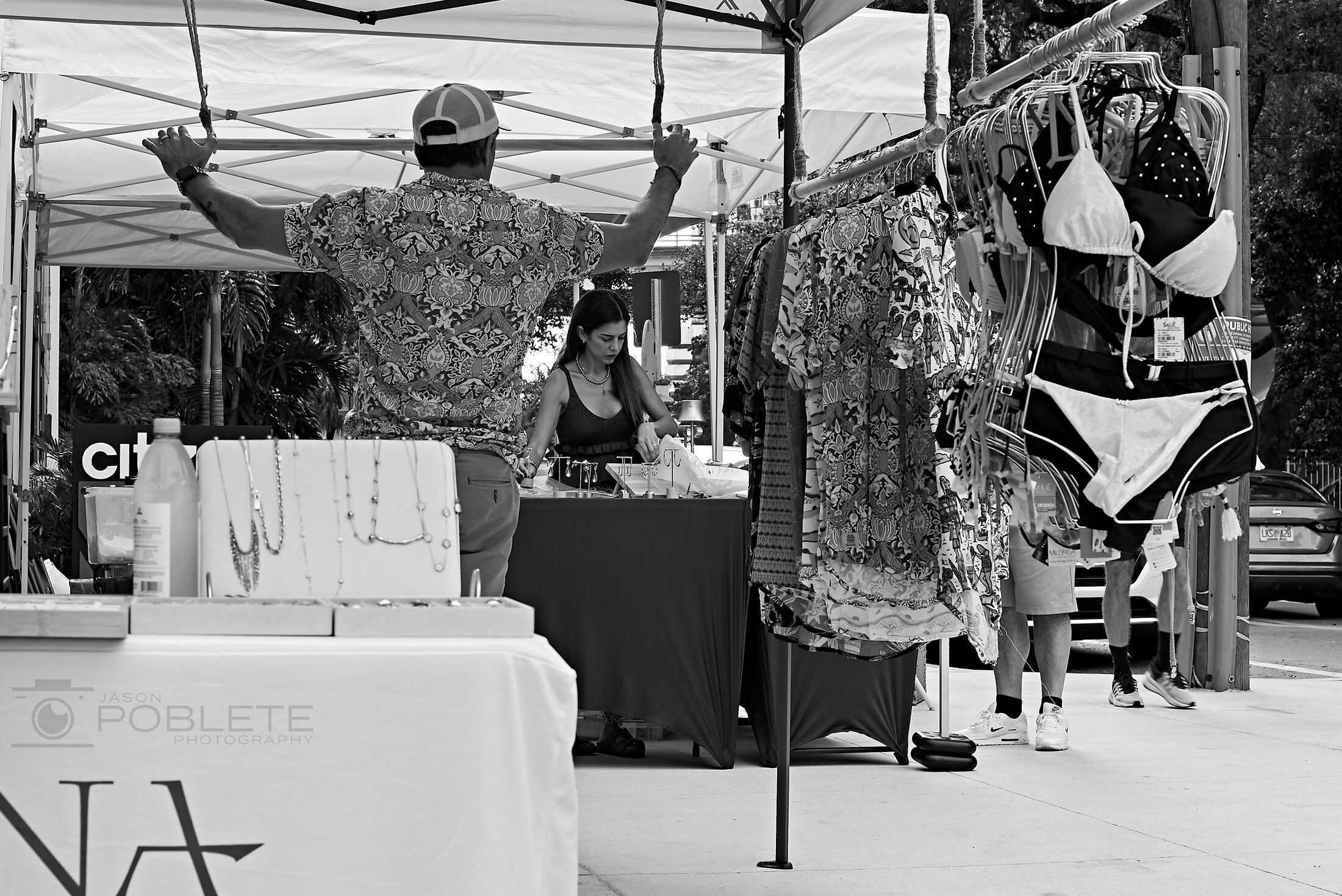 A market vendor stands behind a counter beneath hanging merchandise.
