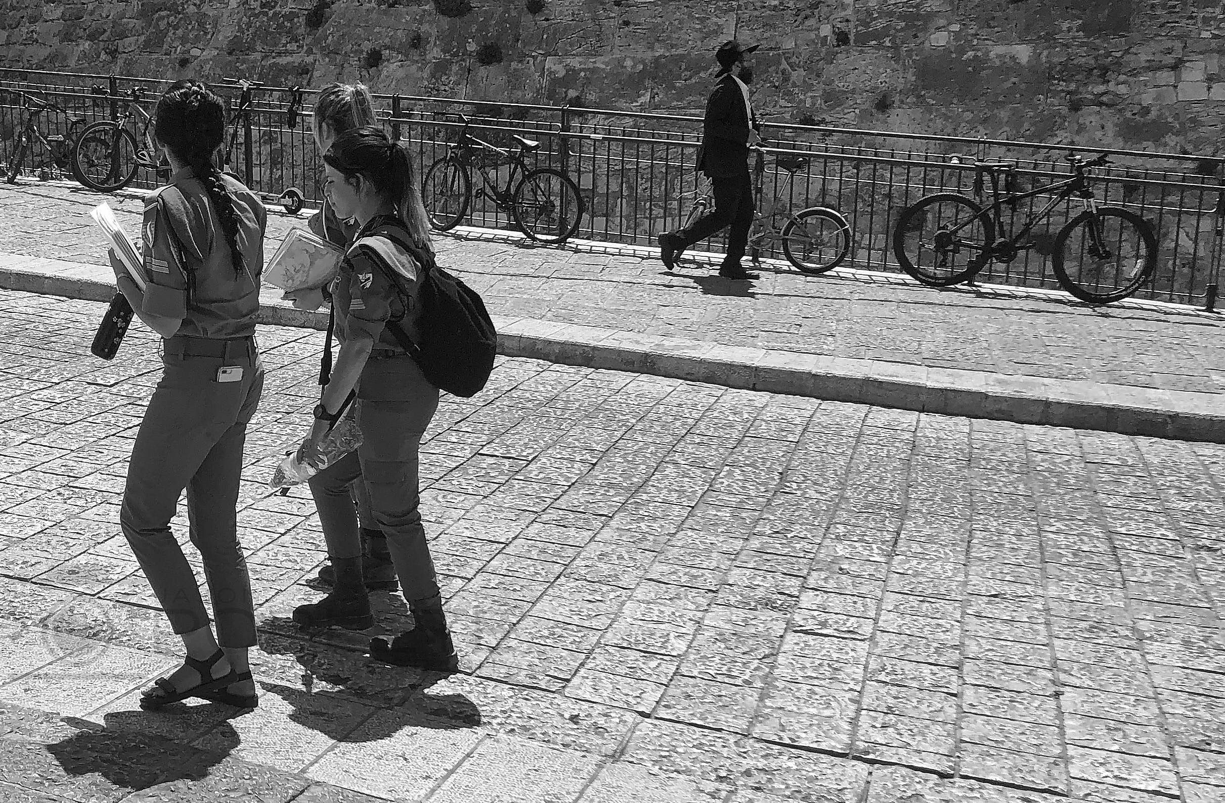 Two women in matching uniforms stand with books on a sunlit stone promenade as a man walks behind them.