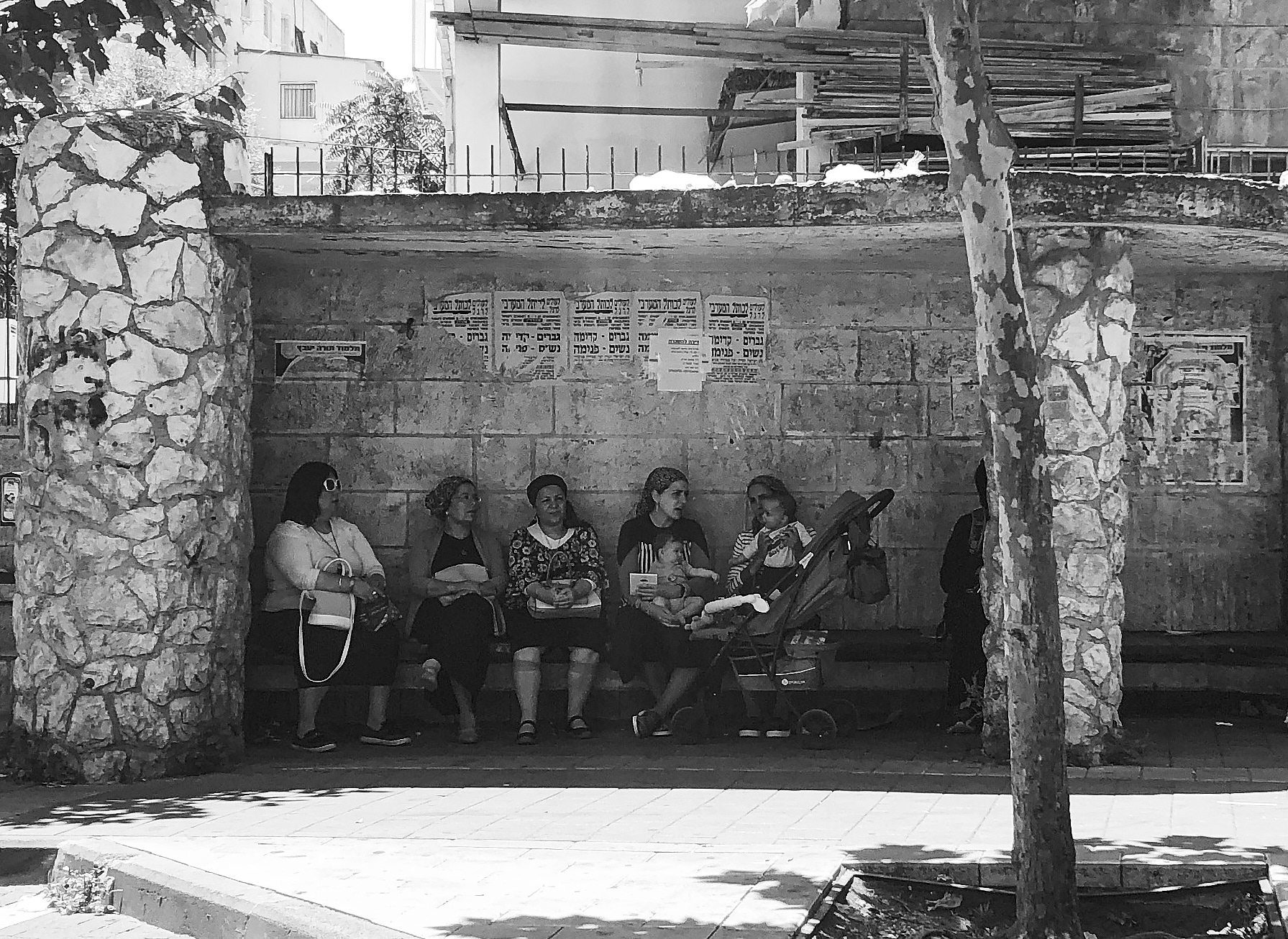 Several women sit together with a stroller in a shaded stone shelter.