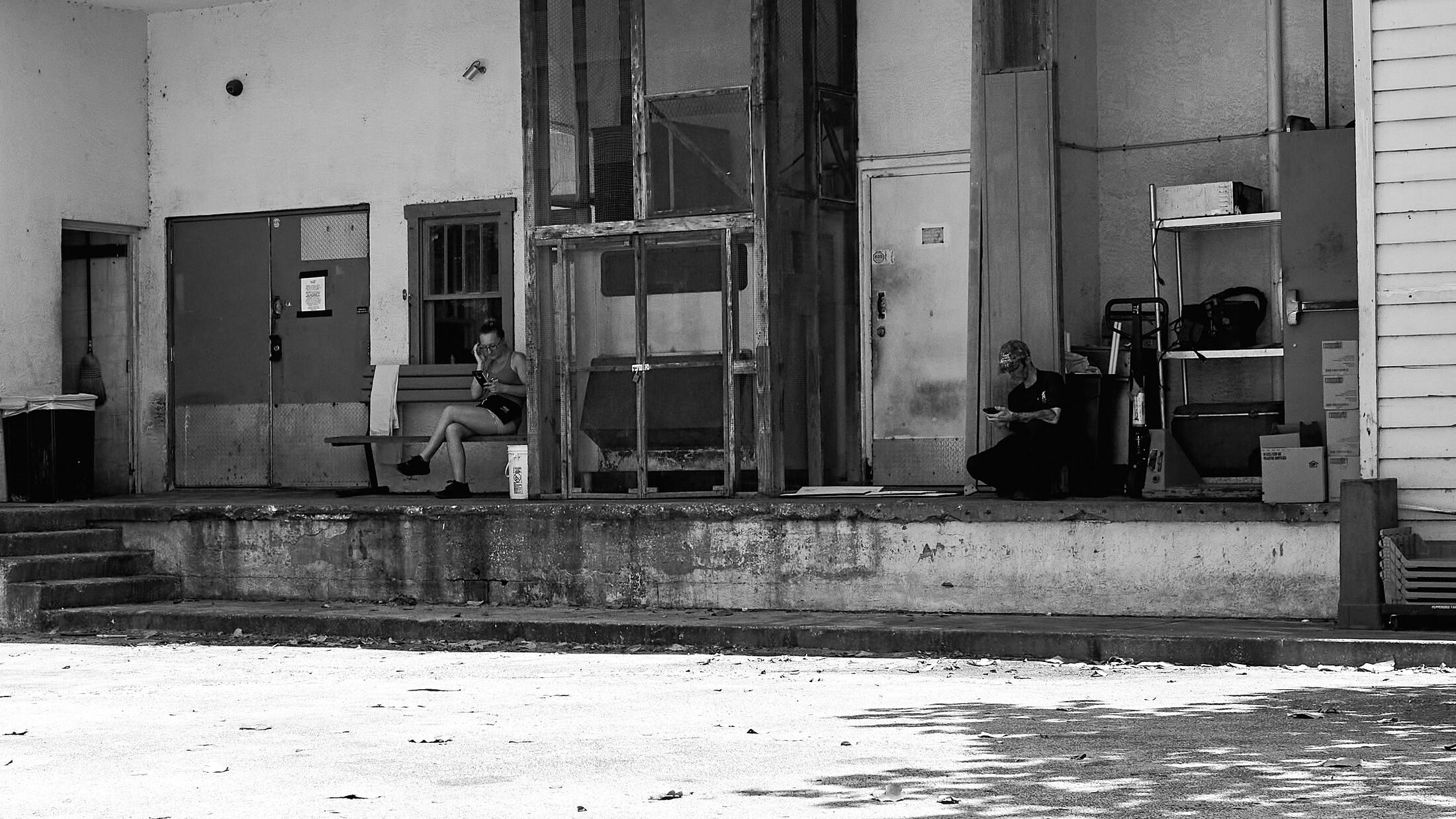 Two people sit apart on a weathered loading dock outside an industrial building.