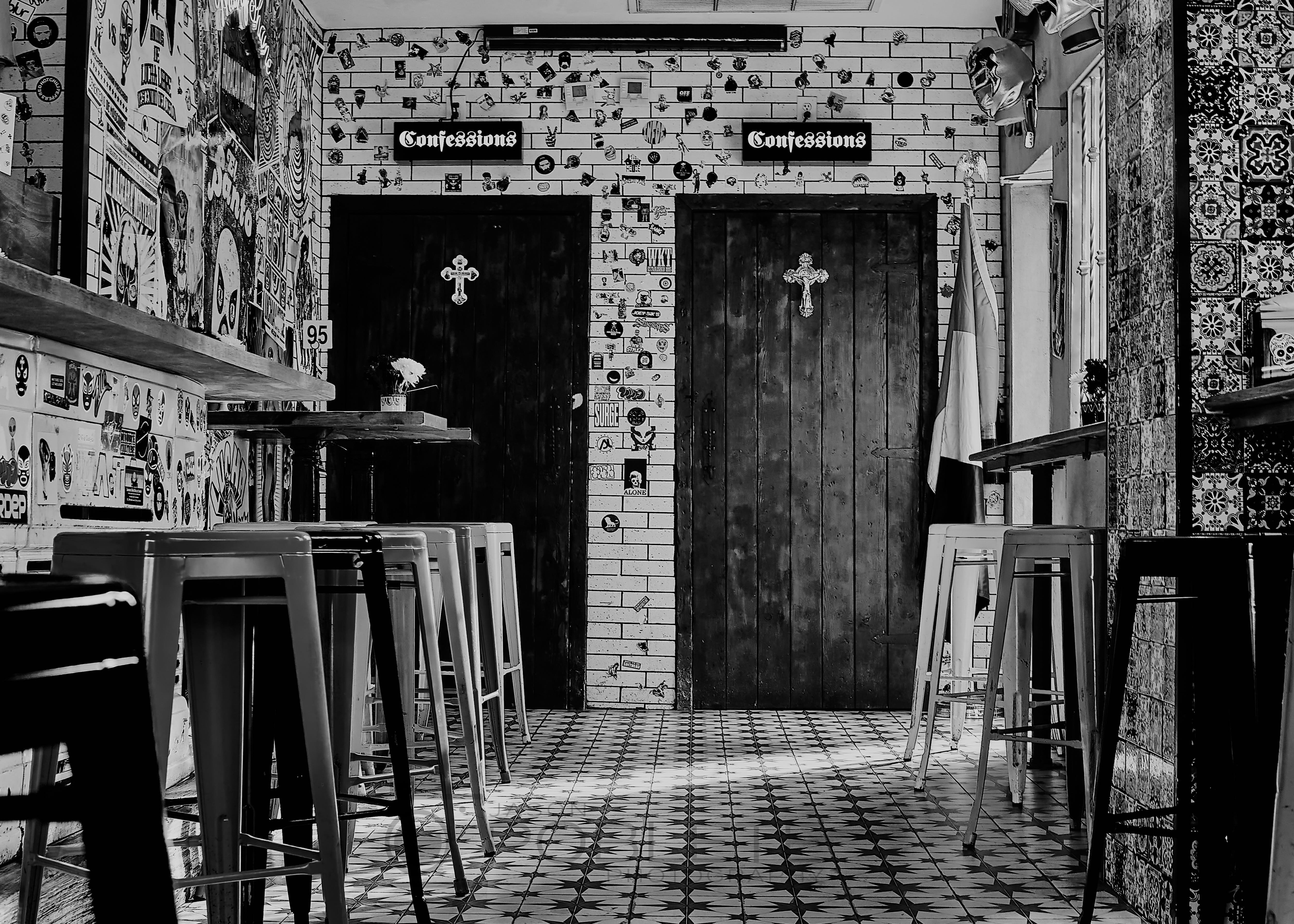 Two dark wooden doors labeled “Confessions” stand inside a tiled room lined with bar stools and signs.