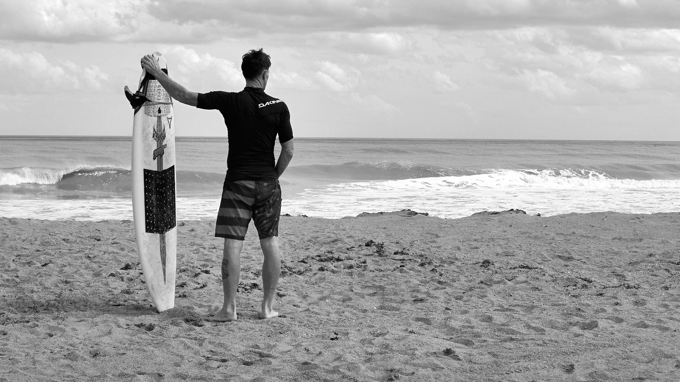 A surfer holds a board upright while looking toward the ocean.
